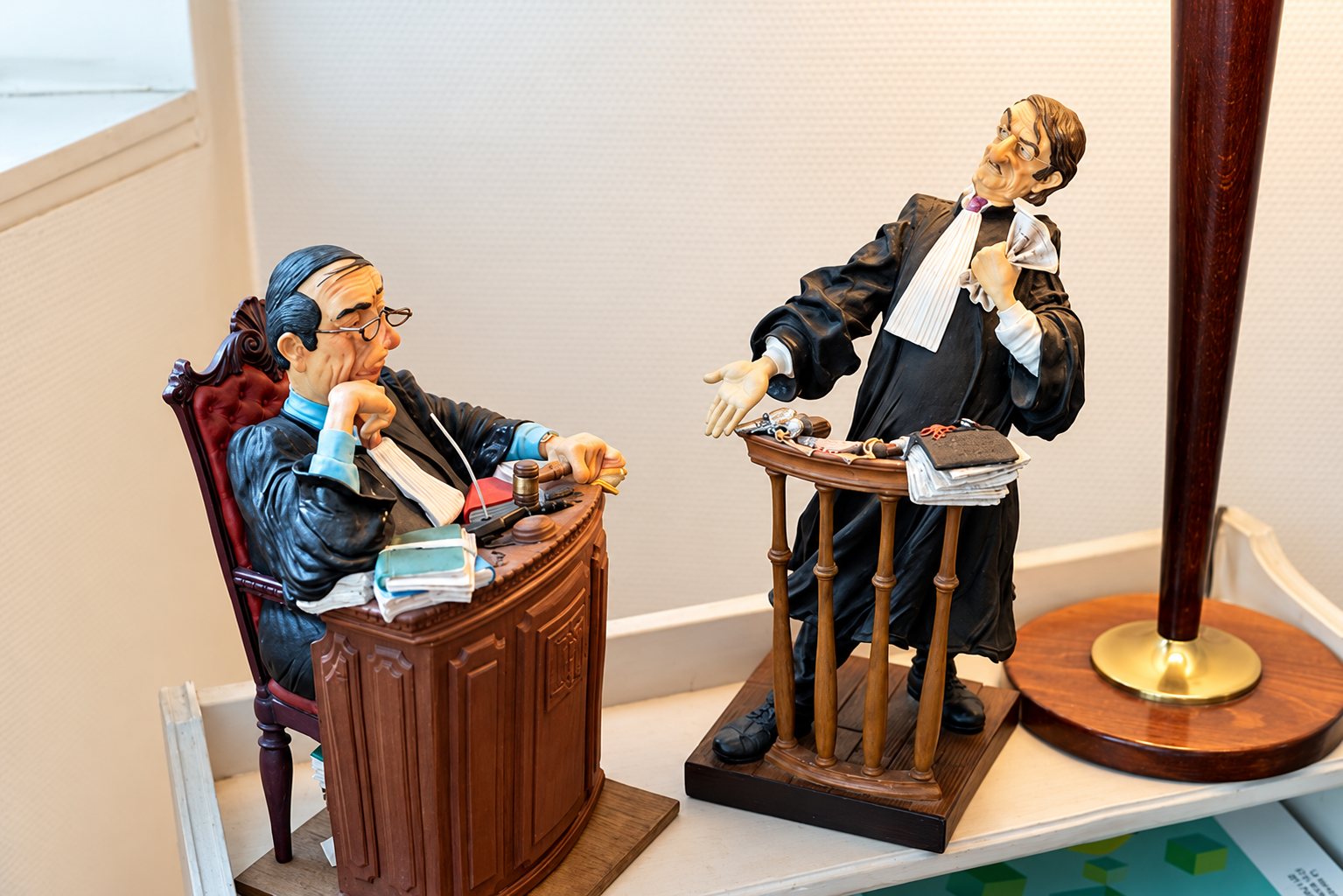 Deux figurines colorées représentant un juge assis derrière son bureau et un avocat en robe plaidant debout, posées sur une étagère près d’un pied de lampe.