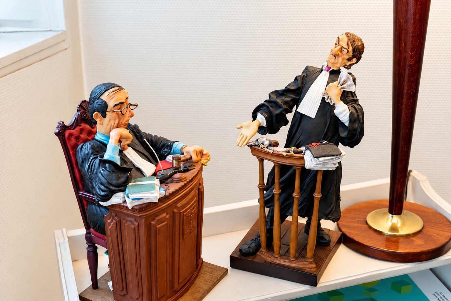 Deux figurines colorées représentant un juge assis derrière son bureau et un avocat en robe plaidant debout, posées sur une étagère près d’un pied de lampe.